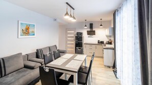 Una sala moderna con un sofá gris, una mesa de comedor con sillas, una zona de cocina con nevera, microondas y armarios, y un cuadro en la pared.
