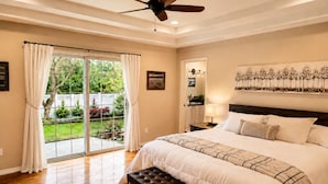 A bedroom with a large bed, a ceiling fan, a view to a garden through a sliding door, and a nightstand with a lamp.