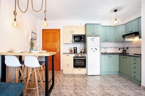 Una cucina con una porta in legno, un forno a microonde, un fornello e un frigorifero. Ci sono due sgabelli e un tavolo.