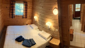 A wooden cabin room with a bed, two wall-mounted lights, a window with curtains, and a door. - Chalet Le Roy in Mauroux – Relaxation, Nature & Family Fun! (Mauroux)
