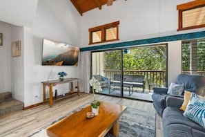A living room with a TV, a coffee table, a sofa, and a balcony with outdoor furniture. - Tropical Treehouse Near the Beach (Koloa)