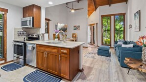 A modern kitchen with stainless steel appliances, wooden cabinets, and a large island. There is a dining area with a blue sofa and a coffee table. - Tropical Treehouse Near the Beach (Koloa)