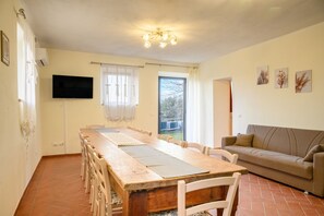 A dining area with a long wooden table, white chairs, a television mounted on the wall, and a sofa in the corner. - Villa Quercina by MMega (Pergine Valdarno)