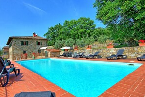 Une piscine avec des chaises longues, un bâtiment en pierre et des arbres en arrière-plan.