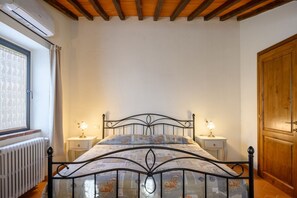 A bedroom with a bed, two bedside tables, a window with curtains, a radiator, and a wooden door.