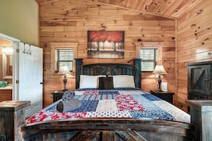 A wooden cabin bedroom with a bed, two bedside lamps, a painting above the bed, and a wooden wardrobe.