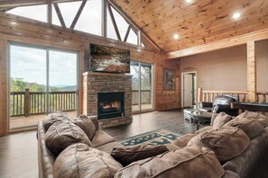 A spacious living room with a stone fireplace, leather sofa, and a large window overlooking a deck. - Beautiful Cabin, Beautiful Mtn Views!! High speed Wifi! Hot Tub, 4 Suites, Great Deals (Sevierville)