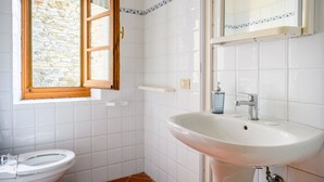 Un bagno con lavandino bianco, un water, una finestra con vista su un muro in pietra e uno specchio sopra il lavandino.