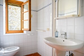 Un baño con un lavabo blanco, un inodoro, un ventanal con vistas a una pared de piedra y un espejo sobre el lavabo.