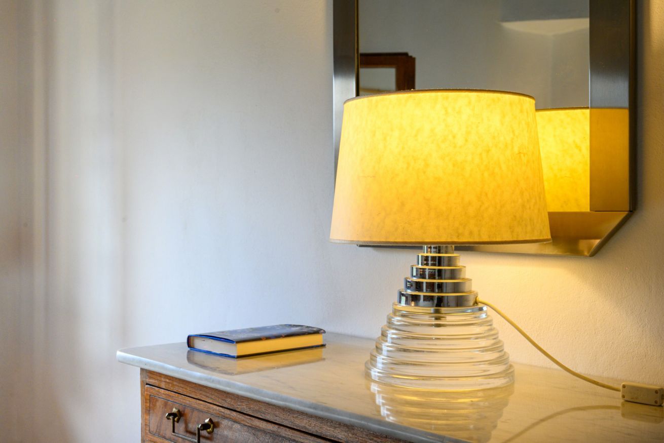 Eine Lampe mit strukturierter Lampenschirm auf einem Nachttisch, ein Buch und ein Spiegel, der die Lampe reflektiert.