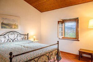 Un dormitorio con cama, mesita de noche, lámpara, ventana con persiana abierta y un banco de madera.