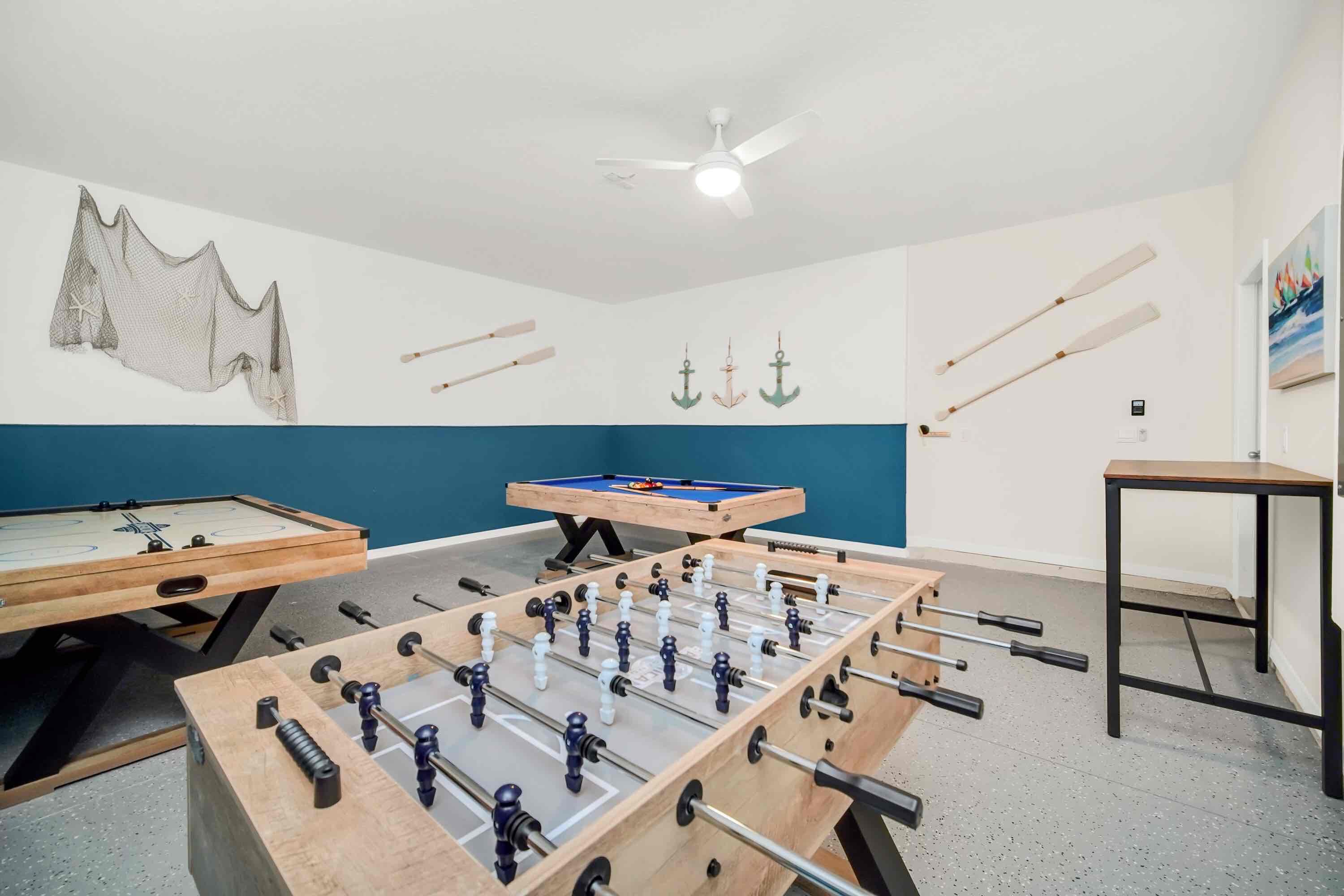 A room with two foosball tables, a pool table, and a ceiling fan.