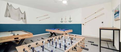A room with two foosball tables, a pool table, and a ceiling fan.