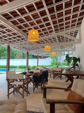 Un espacio exterior para comer con mesas y sillas de madera, lámparas amarillas colgantes y vista a una piscina y vegetación.