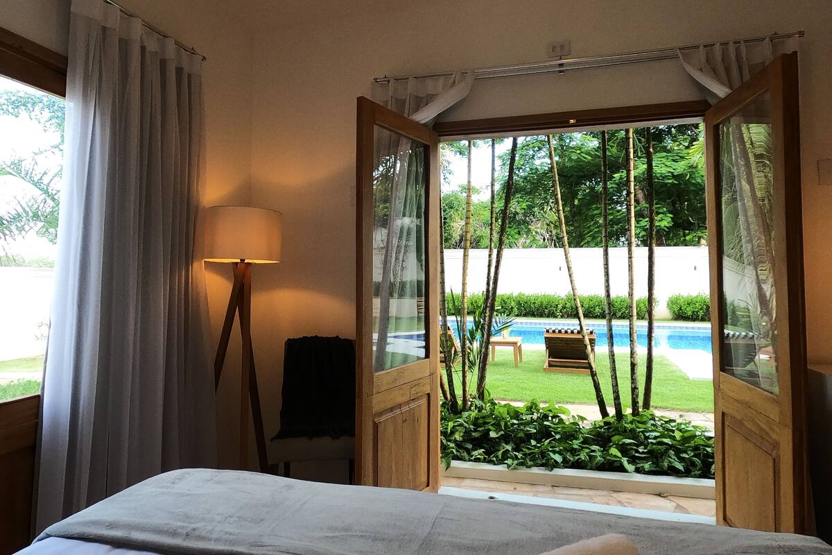Quarto de hotel com cama, luminária e vista para uma piscina.