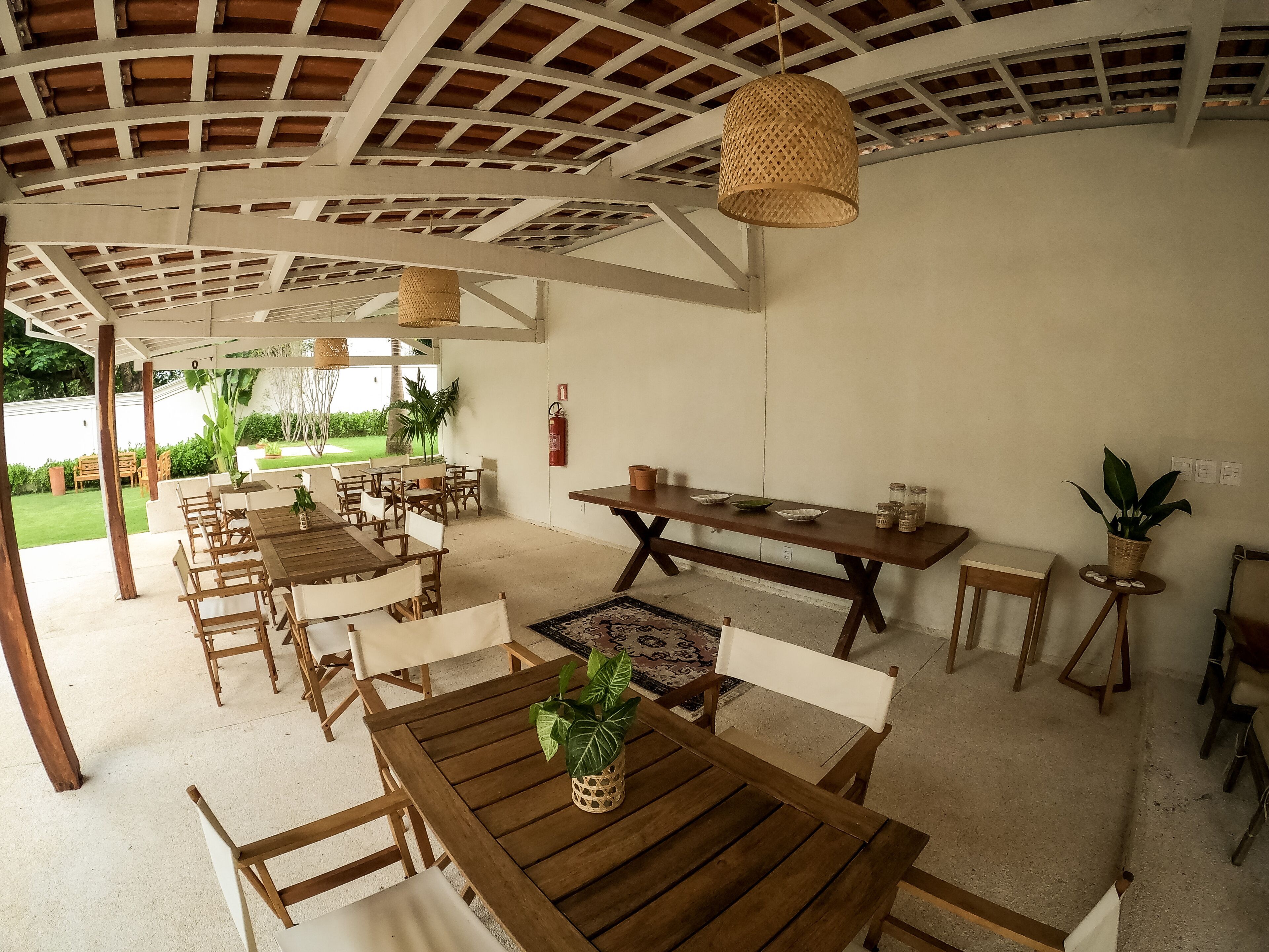 A covered outdoor dining area with wooden tables and chairs, a fire extinguisher on the wall, and a view of the garden.