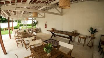A covered outdoor dining area with wooden tables and chairs, a fire extinguisher on the wall, and a view of the garden.