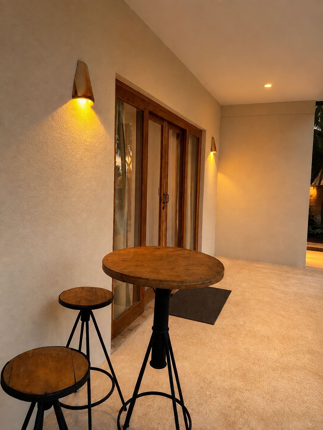 Una habitación con una mesa redonda y dos taburetes, un ventanal corredero y una lámpara fijada a la pared.