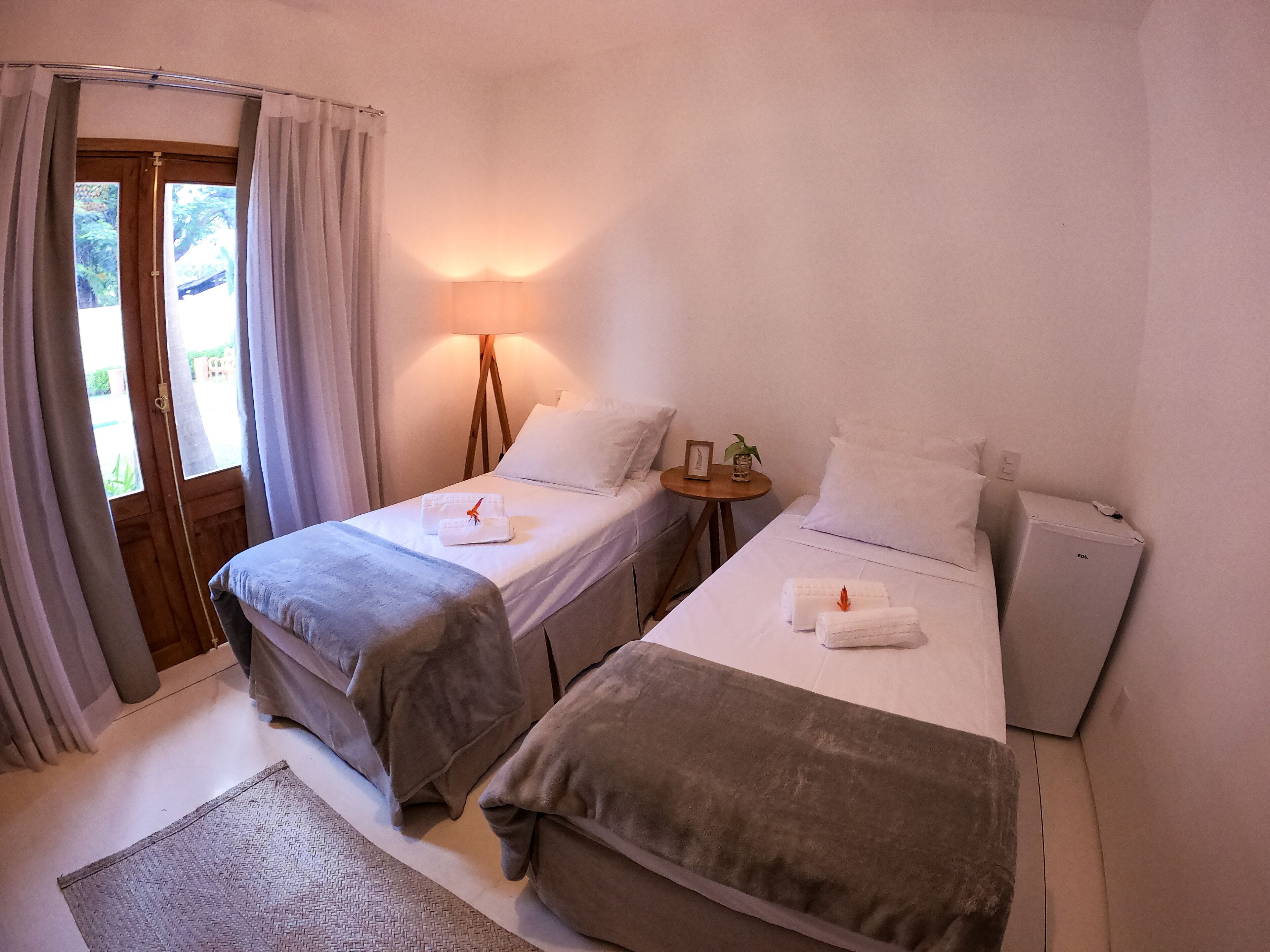 Quarto de hotel com duas camas, uma mesa pequena com abajur e vista pela porta.