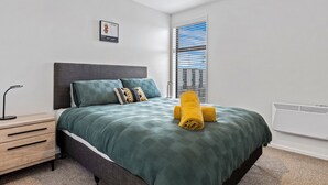 A neatly made bed with a green bedspread, two pillows, and a rolled yellow towel. There is a bedside table with a lamp and a drawer unit. A window with blinds is visible, and a wall-mounted air conditioner is present. - Central Christchurch 2 Bedroom Apartment – Balcony & Parking (Christchurch)