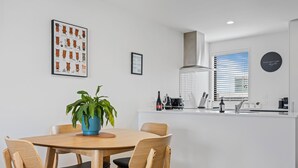 A modern kitchen with a dining area, featuring a round wooden table, black cushioned chairs, a blue plant, and a framed poster on the wall. - Central Christchurch 2 Bedroom Apartment – Balcony & Parking (Christchurch)