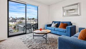 A modern living room with a large sliding glass door, a blue sofa, and a round coffee table. - Central Christchurch 2 Bedroom Apartment – Balcony & Parking (Christchurch)
