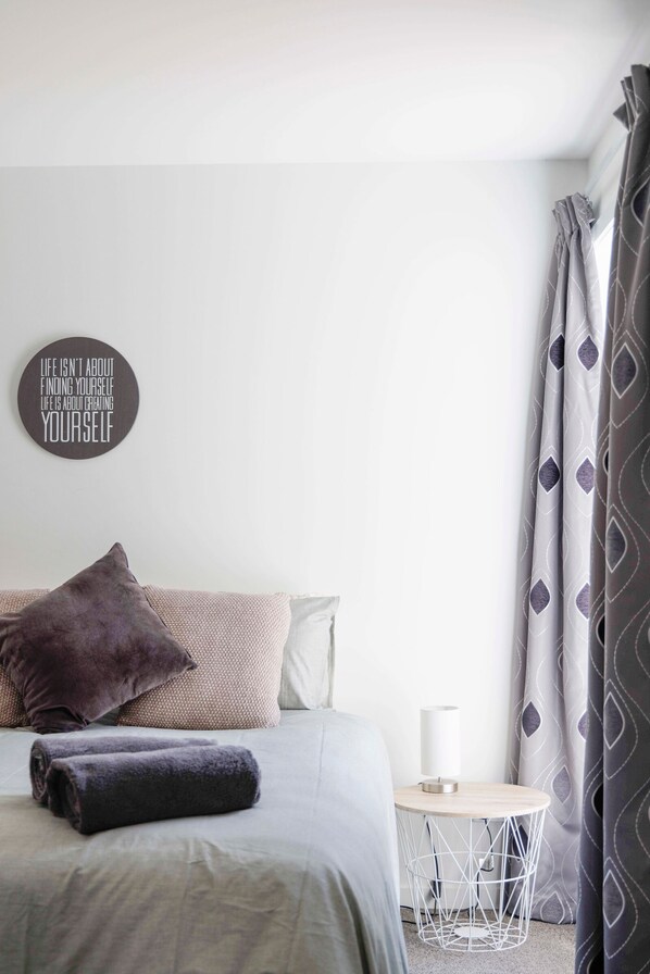 Een keurig opgemaakt slaapkamer met een bed, kussens, een nachtkastje met een lamp en een inspirerende quote op de muur.