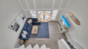 Un vaste espace de vie comprenant un canapé bleu, une table basse et un grand téléviseur. Un escalier mène à un autre niveau.