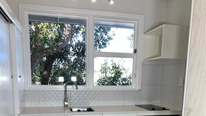 A compact kitchen with white cabinets, a sink, a small refrigerator, and a window with a view of trees. - Kaikōura Peaks Studio Retreat 1 (Kaikoura)