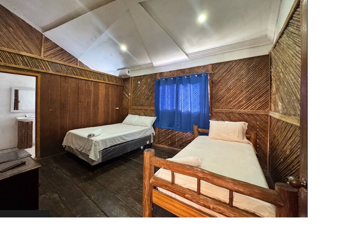 A room with two single beds, a wooden headboard, a small bedside table, and a window with blue curtains.