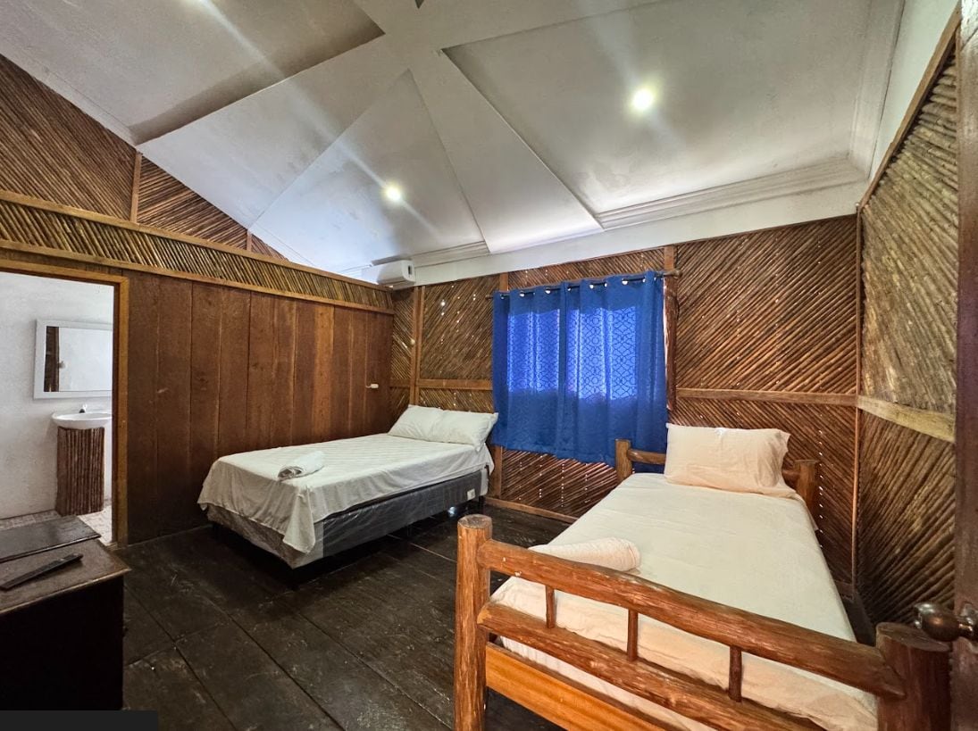 A room with two single beds, a wooden headboard, a small bedside table, and a window with blue curtains.