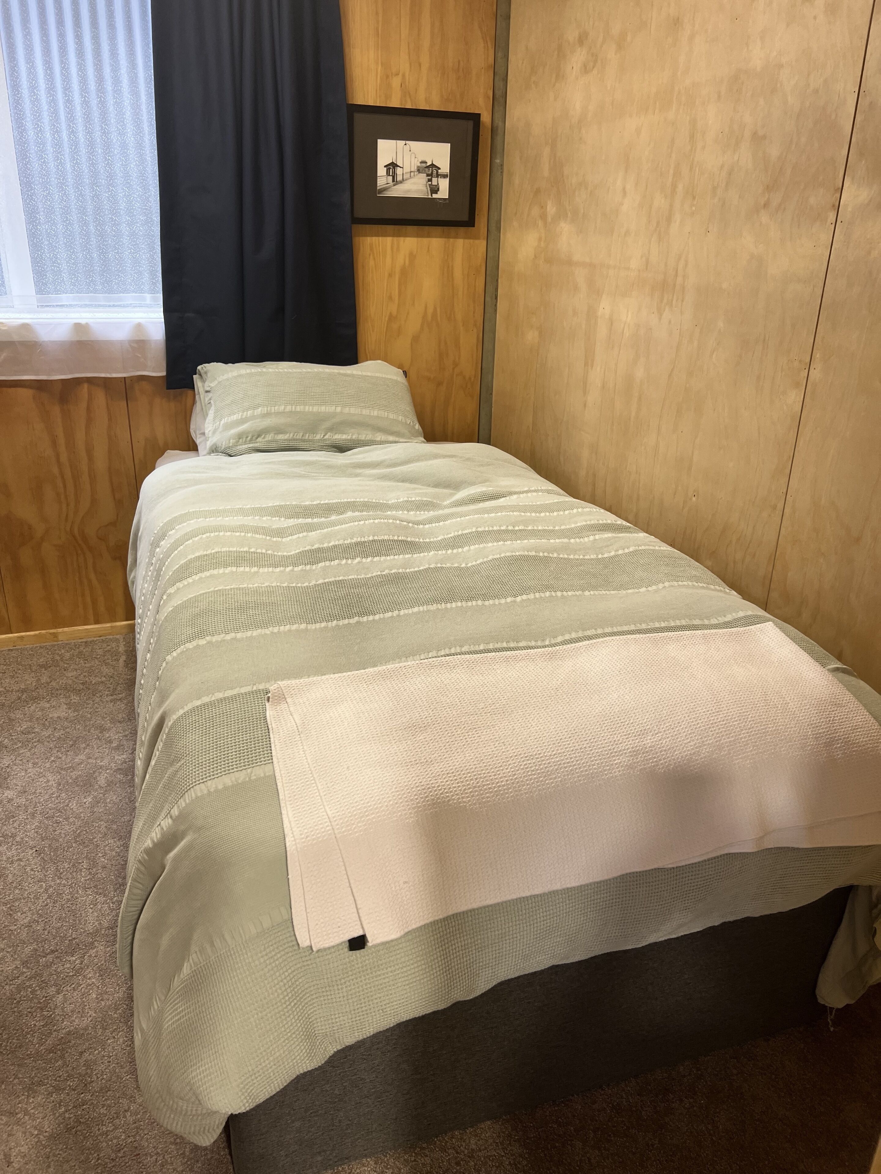 A neatly made bed with a striped bedspread and pillows, a framed picture on the wall, and a window with curtains.