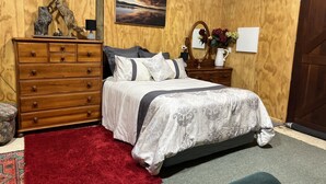 Quarto com cômoda de madeira, cama com colcha estampada, tapete vermelho e pintura de um pôr do sol sobre uma ponte.