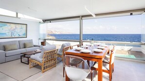 A modern living room with a large window overlooking the sea, a dining area with a table set for four, and a sofa with cushions.