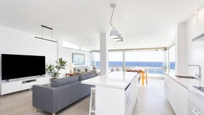 A modern living space with a flat-screen TV, grey sofa, white kitchen, and a view of the sea.