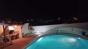 Una piscina amb una estructura de vidre, zona de barbacoa i un petit pati amb taula i cadires.