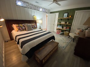 A bedroom with a bed, a wooden bench, a ceiling fan, a nightstand, a lamp, a shelf with items, and a door.