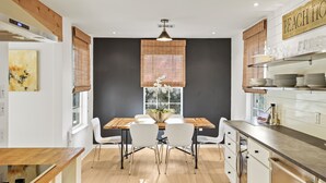 A modern kitchen with a dining area, featuring a wooden table, white chairs, a large window with blinds, and a sign reading "BEACH HOUSE". - Crescent Oasis - Quiet Slice of 30A (Santa Rosa Beach)