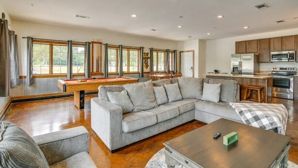 Un salón espacioso con un sofá seccional gris, una mesa de billar y una zona de cocina con electrodomésticos modernos.