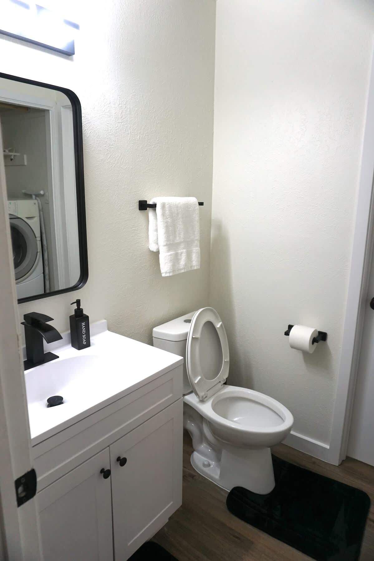 Ein Badezimmer mit weißem Waschbecken, schwarzem Seifenspender, Toilette in Weiß und einer schwarzen Fußmatte.
