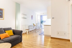 A modern living room with a grey sofa, a dining area with a glass table, and a kitchen in the background.
