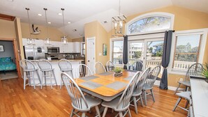 A spacious dining area with a wooden table and multiple chairs, a kitchen with stainless steel appliances, and a large window with a view of the outside.