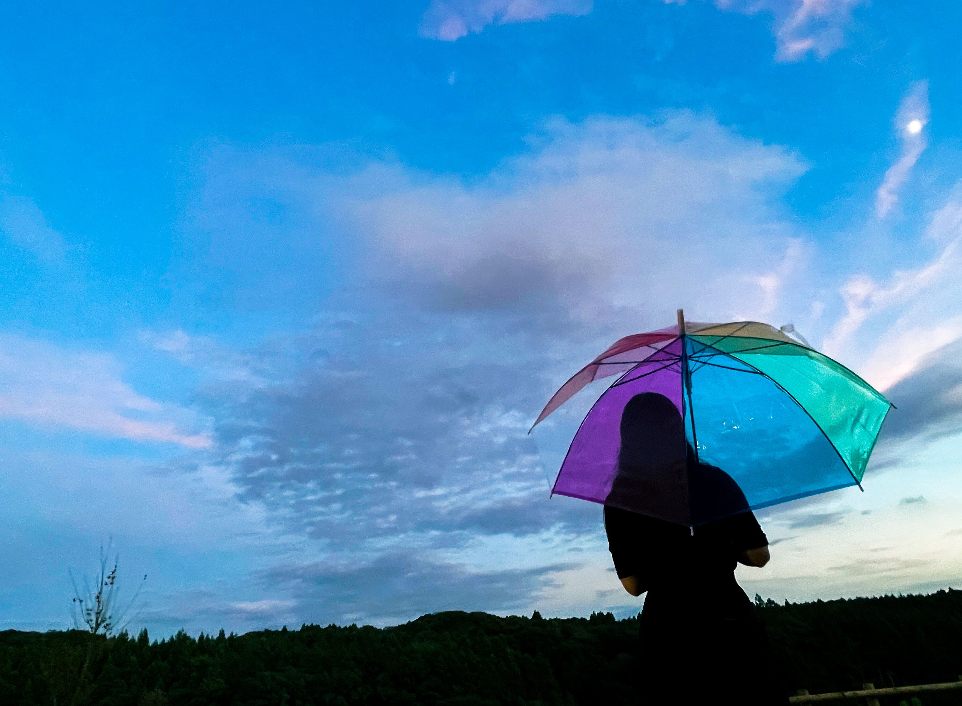 A person holding a transparent umbrella with a visible rainbow pattern, standing against a backdrop of a clear sky with some clouds.