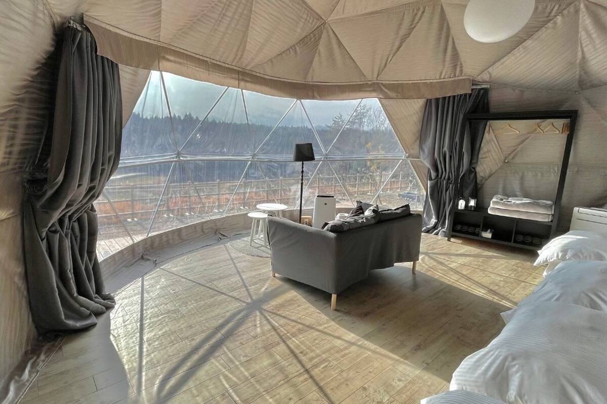 Quarto espaçoso com uma grande janela que oferece vista para uma área arborizada. Há um sofá, uma mesa pequena e uma cama com roupa de cama branca.
