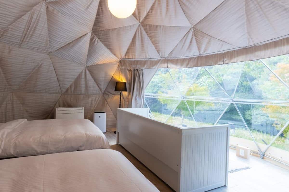 Quarto tipo tenda com cama, mesa de cabeceira, luminária e vista para árvores através de uma grande janela.