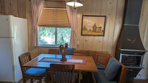 Un coin repas chaleureux avec des murs en bois, une table, des chaises, un banc, une fenêtre avec des stores, un luminaire au plafond et une cheminée.