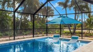 Piscina com duas espreguiçadeiras e um guarda-sol azul, cercada por um espaço envidraçado onde se veem palmeiras.