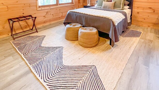 A wooden cabin bedroom with a bed, a nightstand, a mirror, a window, and a wall decoration. - Cute 3-3 cabin with fire pit, games, resort amenities! (Ellijay)