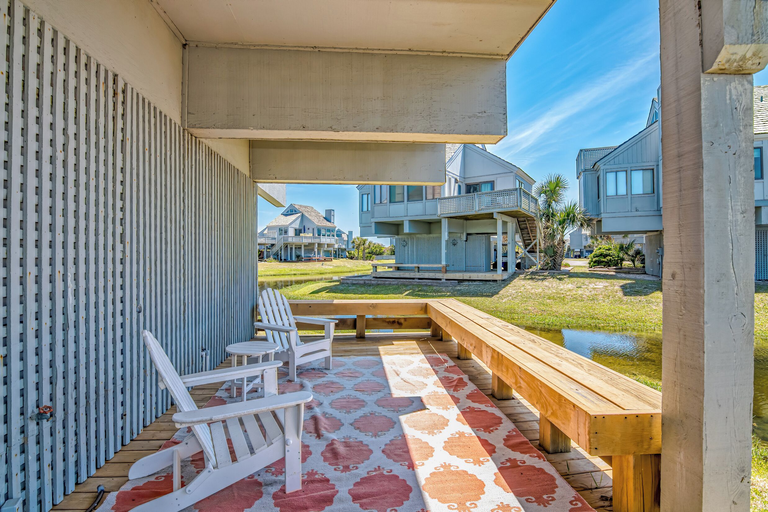A covered outdoor area with a wooden deck, two white Adirondack chairs, and a patterned rug.