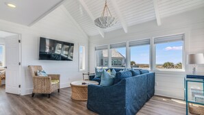 Een ruime woonkamer met een grote blauwe hoekbank, een rieten stoel, een rond rieten bijzettafeltje en een flatscreen televisie die aan de muur is bevestigd.